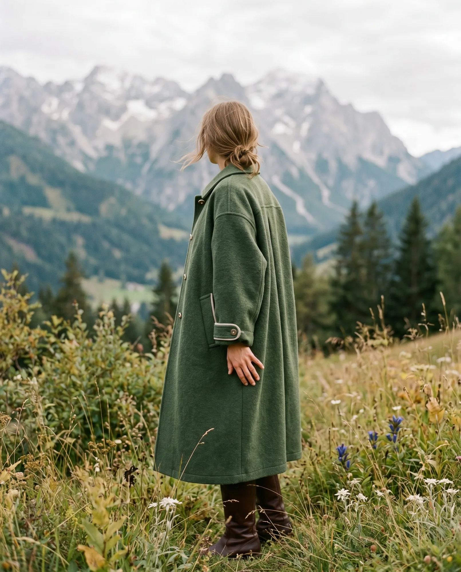 A woman wearing a modern reinterpretation of a traditional Tyrolean loden wool coat in an alpine meadow.
